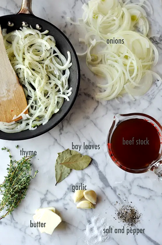 An aerial shot of skillet with onions in it and ingredients on a table including: onions, thyme, bay leaves, garlic, butter, beef stock, salt and pepper, and garlic