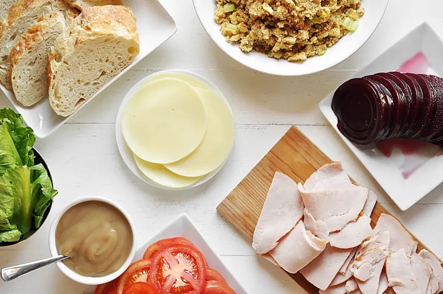 A close up of a a plate of bread, a plate of cheeses, a plate of cranberries, a cutting board of turkey slices, and a plate of tomatoes