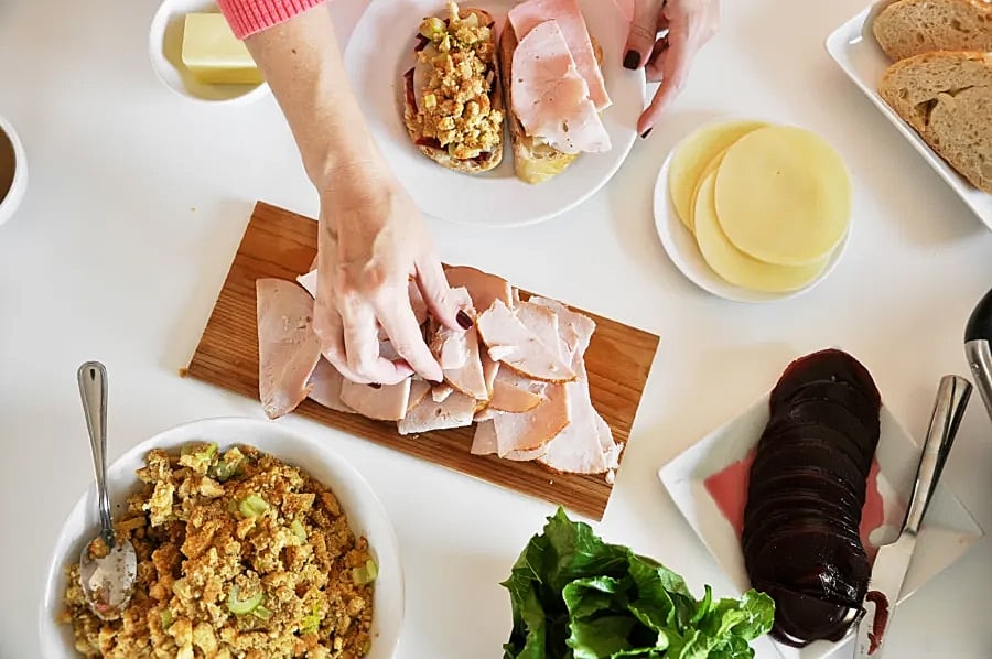 A close up of a woman's hand grabbing a slice of turkey from a cutting board next to a plate of cheeses, a plate of fresh bread, a bowl of stuffing, and a plate of canned cranberries