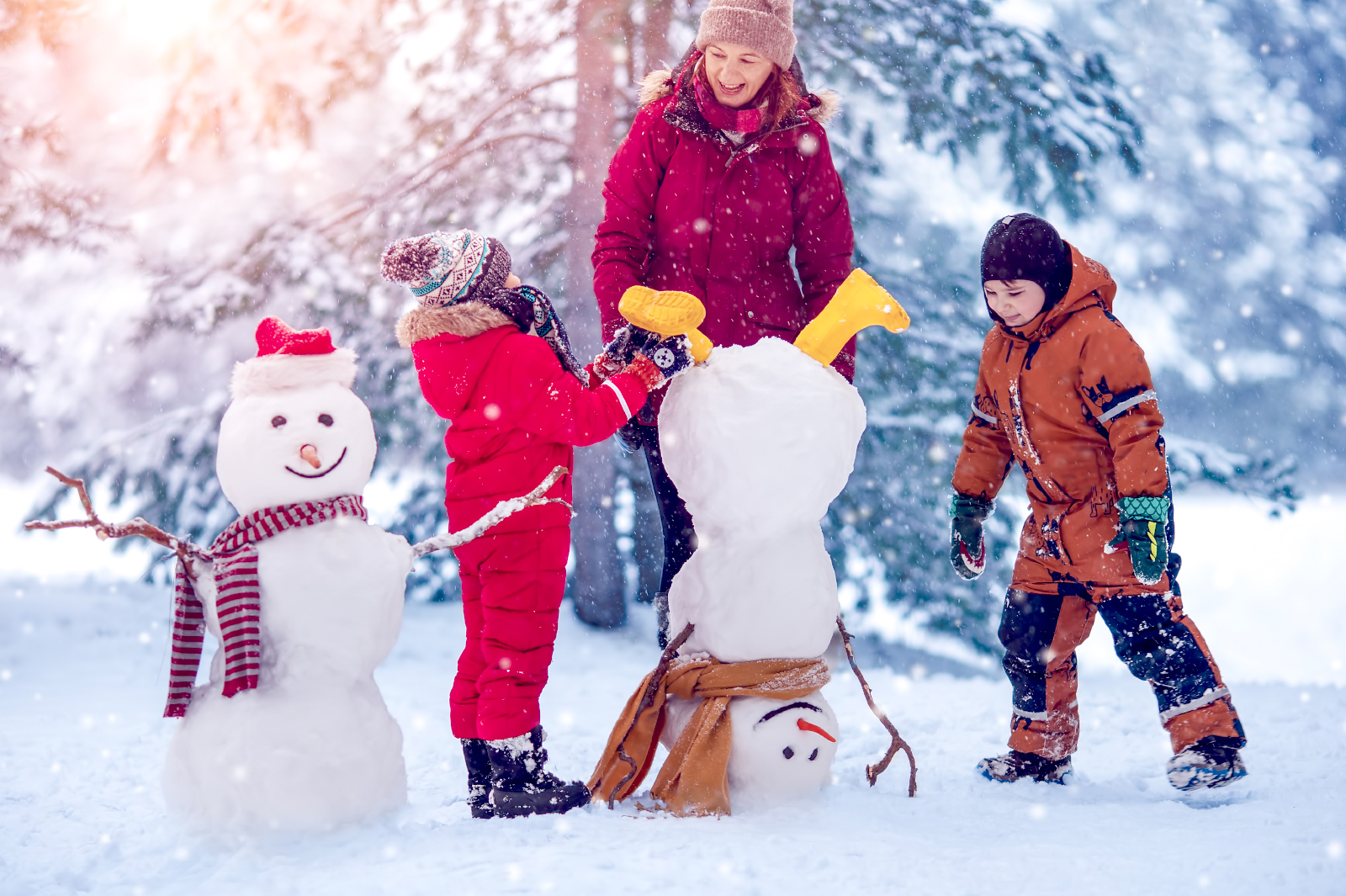 A mom and her two kids playing in the snow, building two snowmen. One is upside down and the other is right side up.