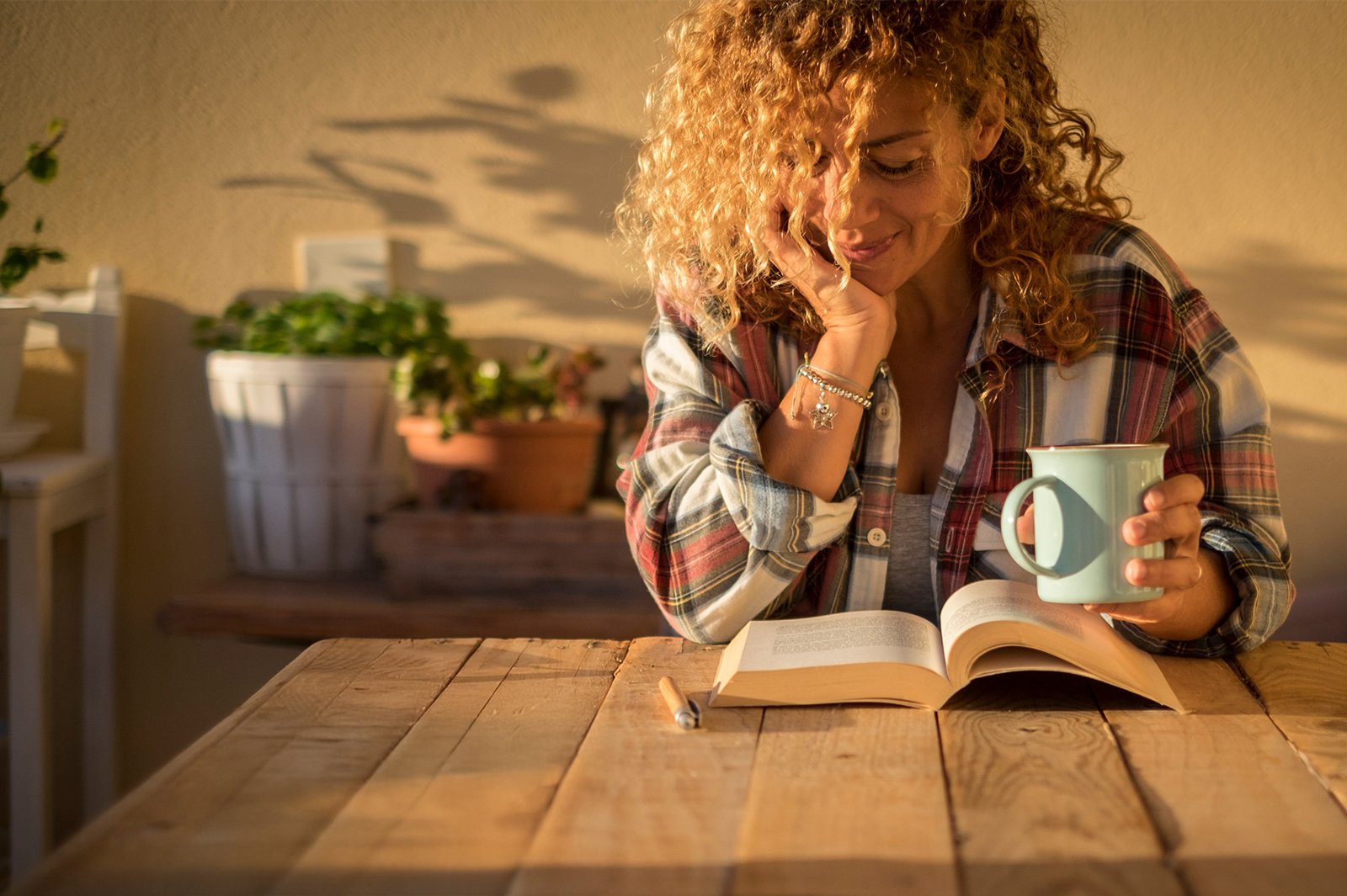 A woman reading a book and holding a cup of coffee as the sunlight pours through the window